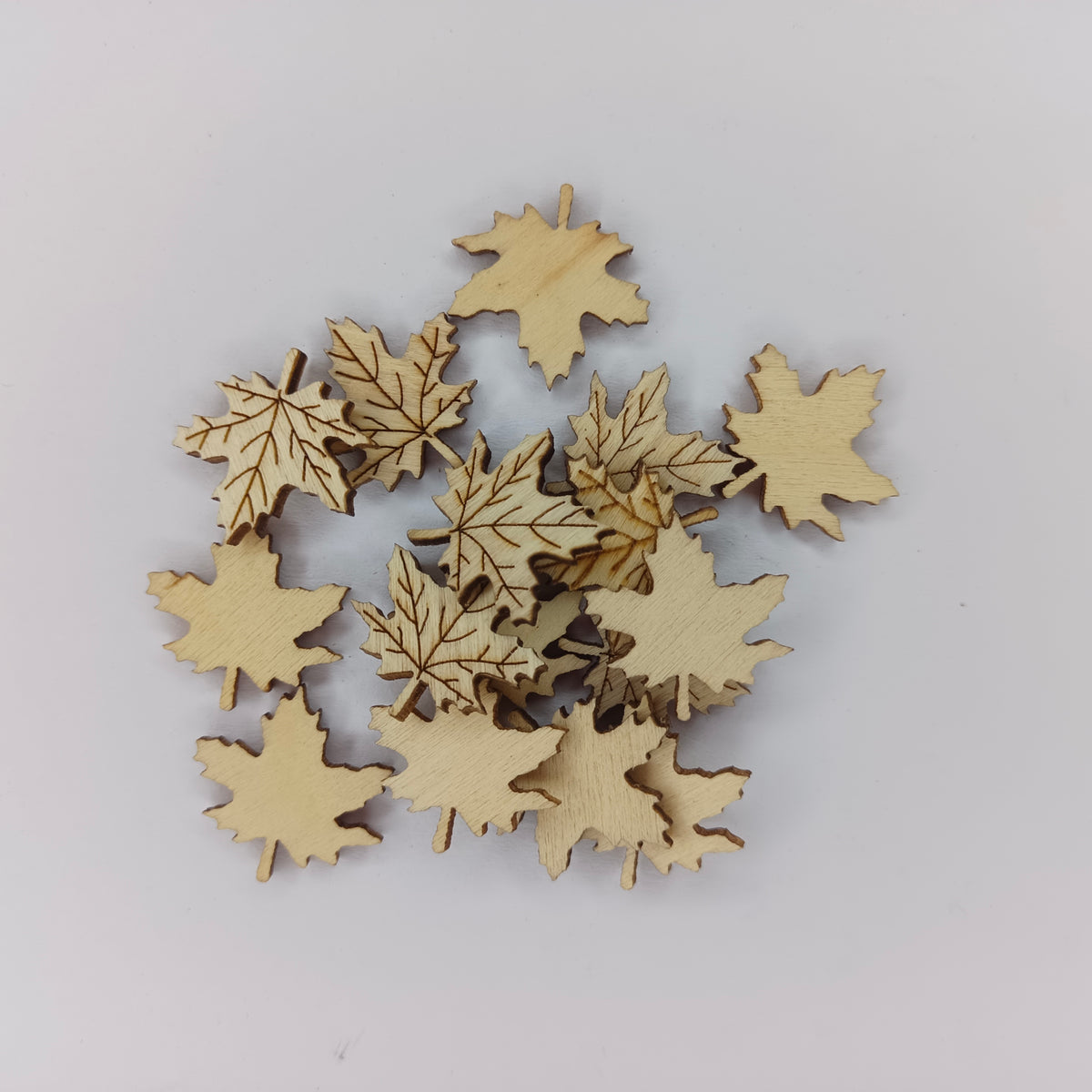 A pile of small laser-cut wooden maple leaves in natural light wood, arranged on a plain light gray background.