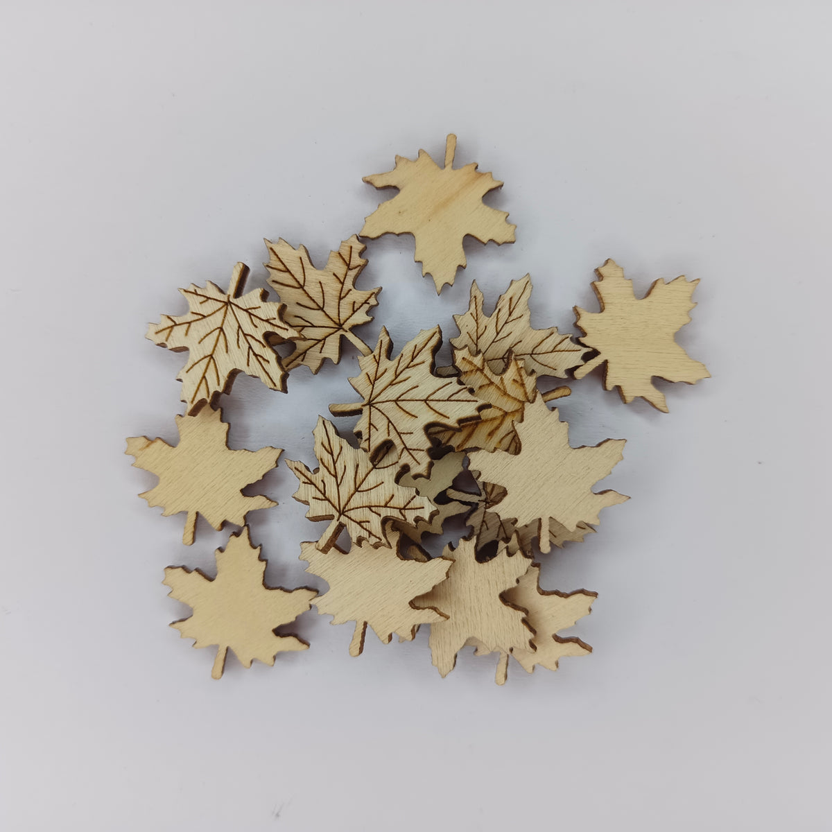 A pile of small laser-cut maple leaf embellishments made of natural light wood, arranged on a plain light gray background.