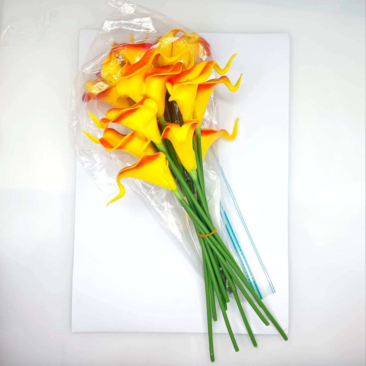 A bouquet of artificial yellow calla lilies with orange-red tips and green stems, wrapped in clear cellophane and placed on a white sheet of paper.