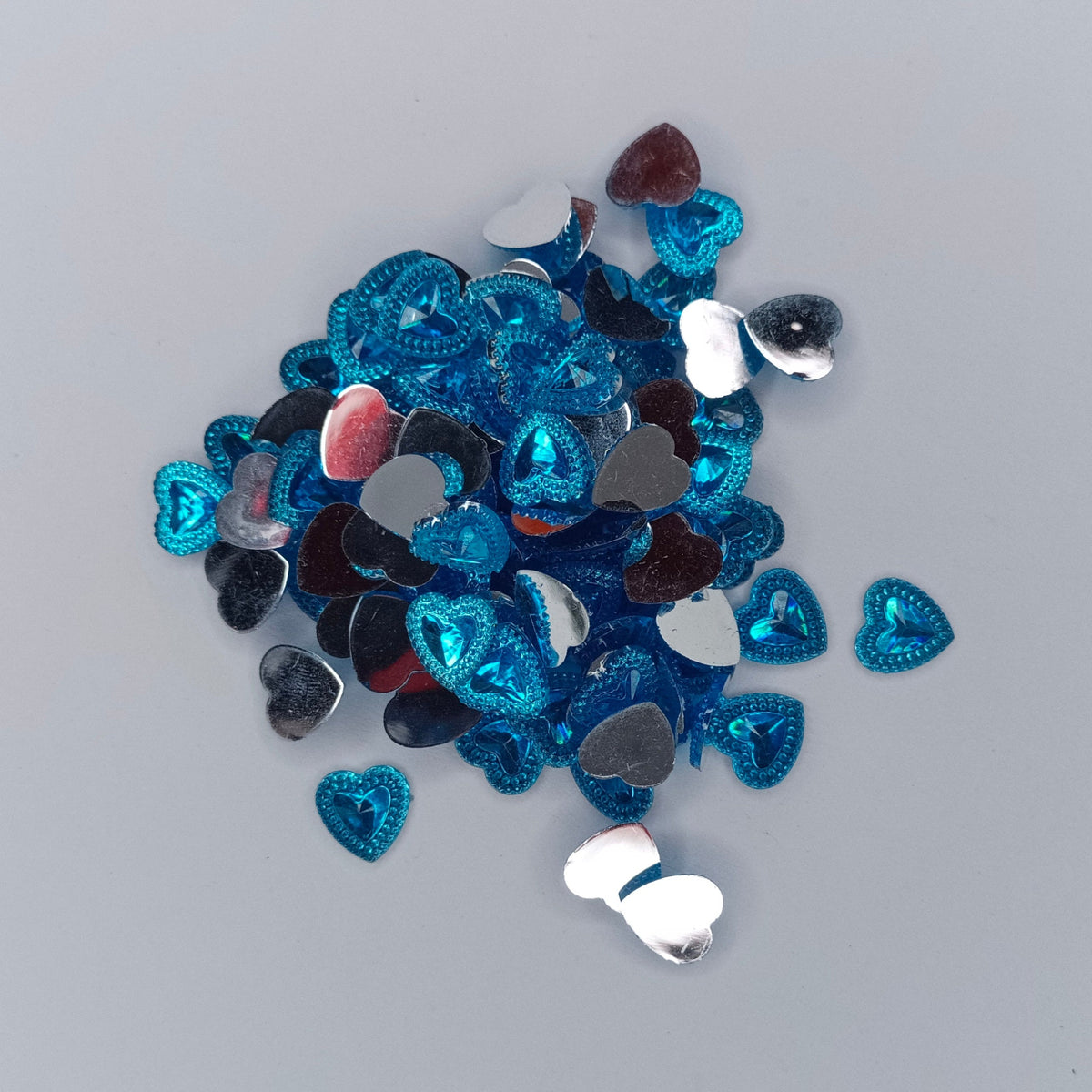 A pile of small heart-shaped rhinestone embellishments in shades of blue, silver, and white, scattered on a plain light gray background.