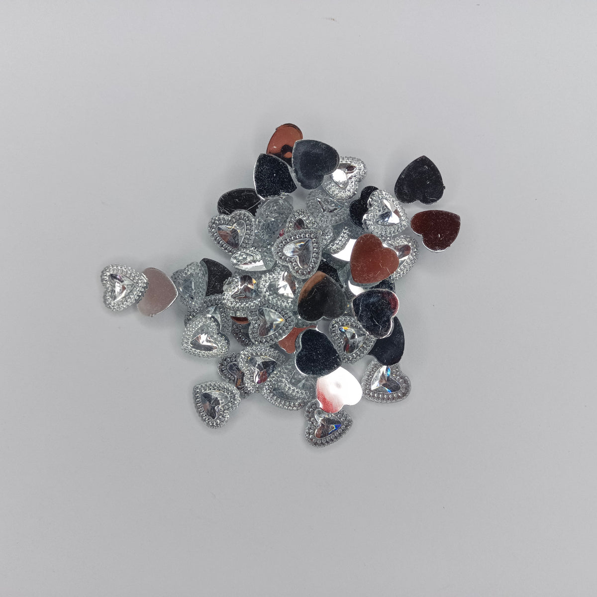 A pile of small heart-shaped rhinestones in silver, black, and reddish-brown, arranged on a plain light gray background.