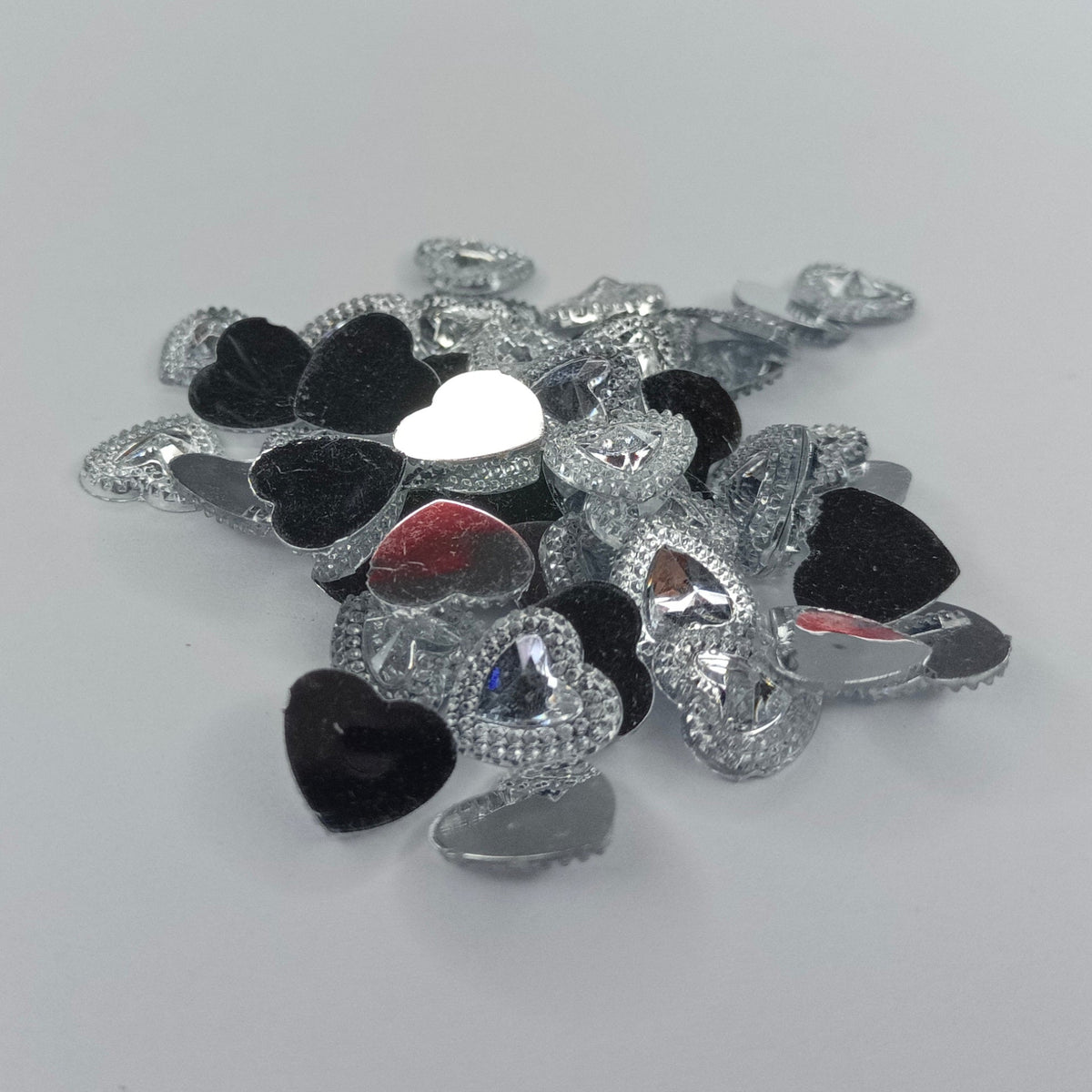 A small pile of heart-shaped rhinestone embellishments in silver, black, and white, photographed on a plain light gray surface.