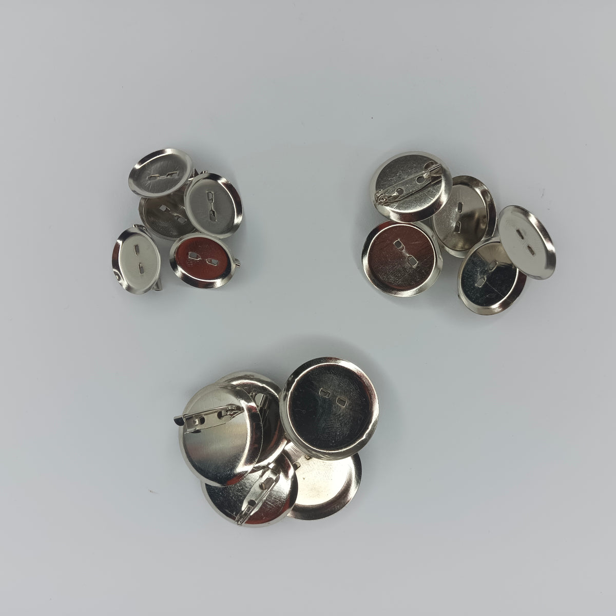 Several small round metallic pin-back cabochon holders, mostly shown from the back on a light gray background. The buttons have silver-toned metal backs.
