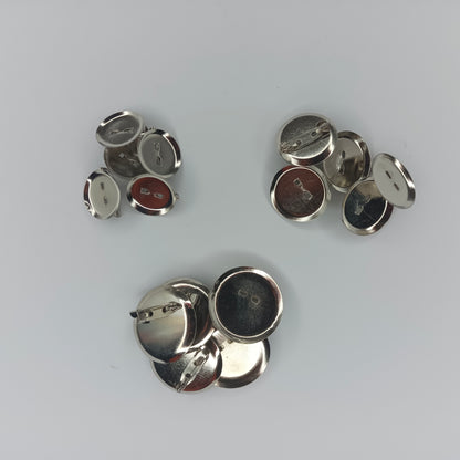 Several small round metallic pin-back cabochon holders, mostly shown from the back on a light gray background. The buttons have silver-toned metal backs.