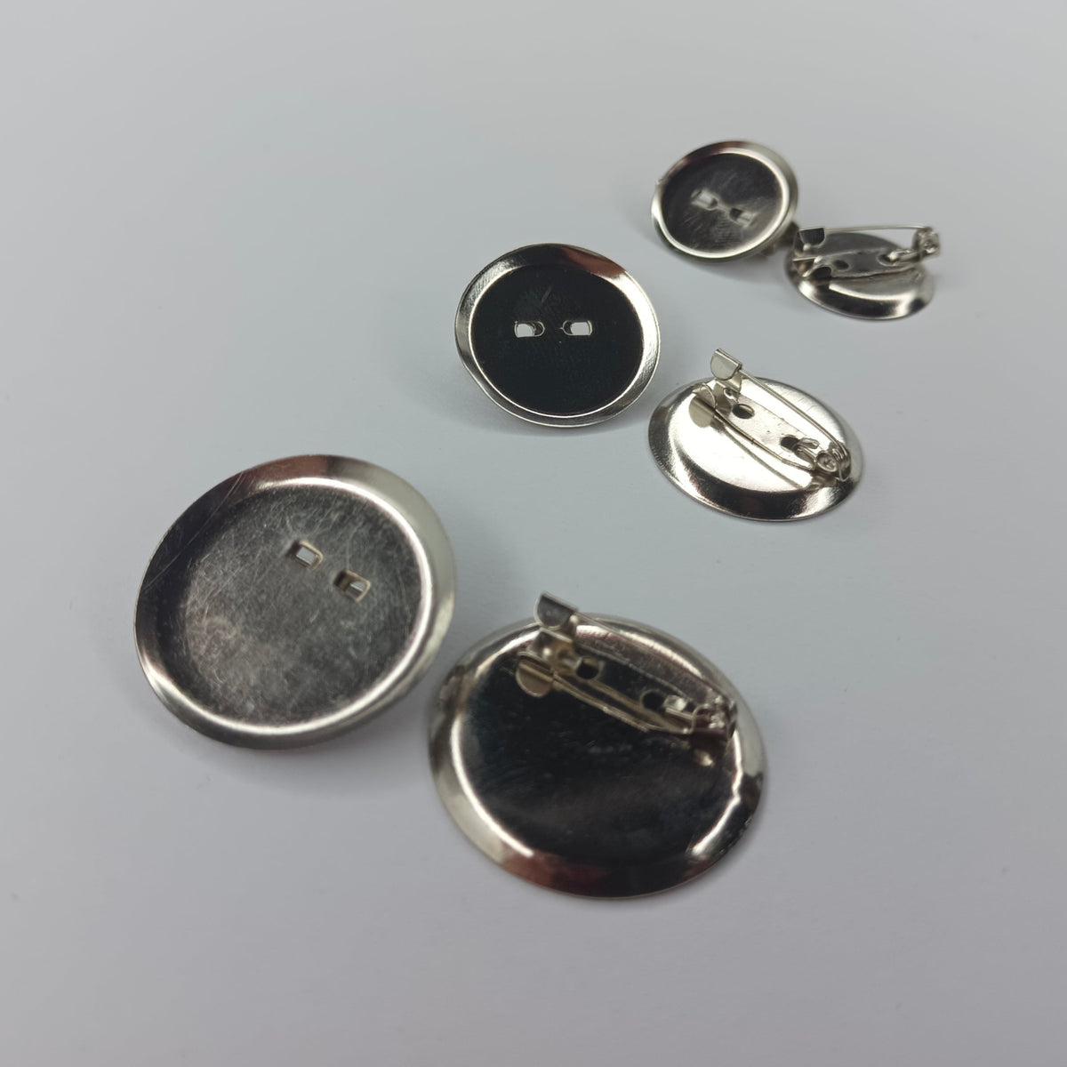 Several small round metallic pin-back cabochon holders, mostly shown from the back on a light gray background. The buttons have silver-toned metal backs.