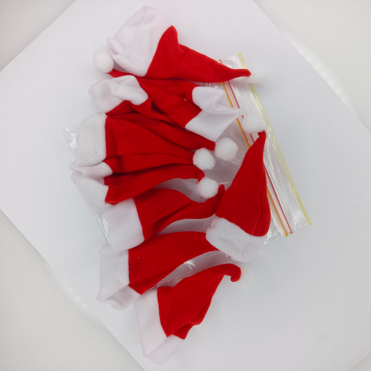 Several small red and white Santa hat felt pieces with white pom-poms are arranged on a white surface next to a clear plastic zip bag.