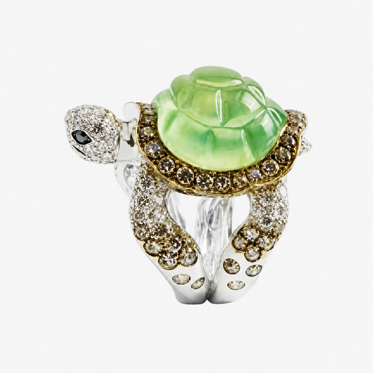 An intricate turtle-shaped ring featuring a large light green carved stone on the shell, a silver-toned body paved with clear stones, and gold-toned detailing around the shell and head.